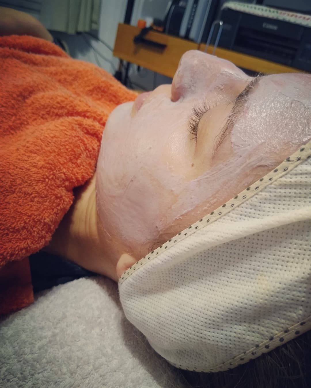 Una clienta recostada en la camilla con una máscara facial lila, vincha blanca y toalla naranja